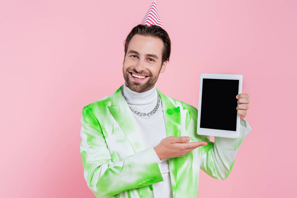 Veselý muž v čepici a bundě ukazuje na digitální tabletu izolovanou na růžové  - Fotografie, Obrázek