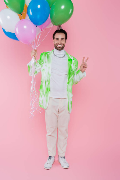 Cheerful man in party cap showing peace sign and holding balloons on pink background  - Valokuva, kuva