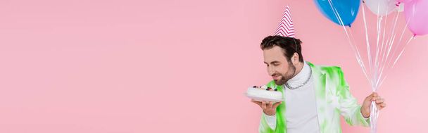 Hombre positivo en gorra de fiesta oliendo pastel de cumpleaños y globos aislados en rosa, pancarta  - Foto, imagen