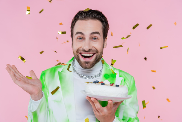 Cheerful man holding birthday cake under confetti on pink background  - Foto, Imagem