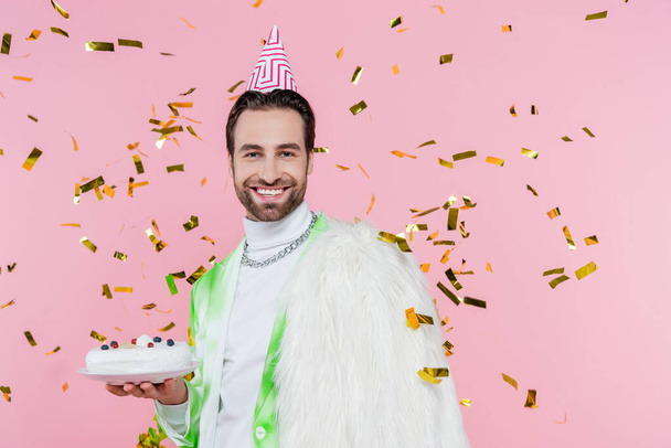 Positive man in furry jacket and party cap holding birthday cake under confetti on pink background  - Фото, изображение