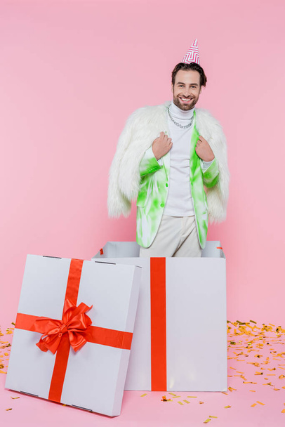 Hombre positivo en gorra de fiesta y chaqueta peluda mirando a la cámara cerca de la caja de regalo enorme y confeti sobre fondo rosa  - Foto, imagen