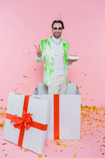 Hombre feliz en gorra de fiesta y gafas de sol mostrando signo de paz y celebración de pastel de cumpleaños en caja de regalo cerca de confeti en rosa - Foto, imagen