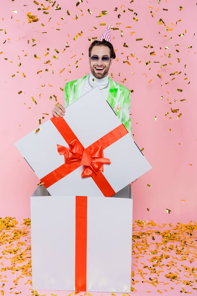 Homem sorridente em óculos de sol e boné de festa em pé na caixa de presente sob confete em rosa  - Foto, Imagem