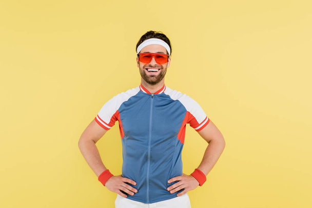Cheerful sportsman in sunglasses holding hands on hips isolated on yellow  - Valokuva, kuva