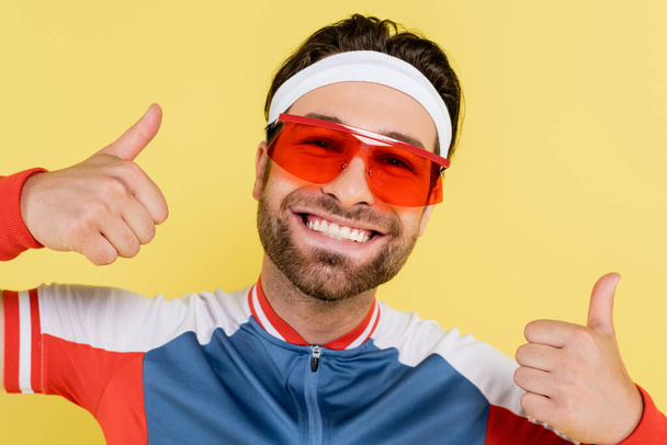 Happy sportsman in sunglasses showing thumbs up isolated on yellow  - Photo, Image