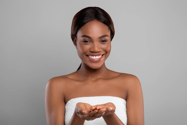 Счастливая черная женщина, завернутая в банное полотенце и демонстрирующая что-то на пустых ладонях - Фото, изображение
