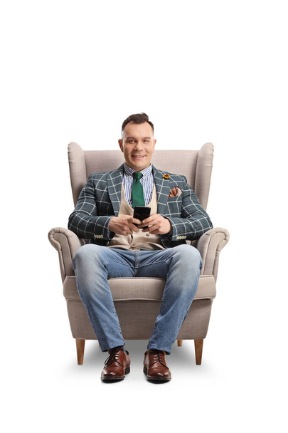 Muž sedí v křesle, drží smartphone a dívá se na kameru izolovanou na bílém pozadí - Fotografie, Obrázek
