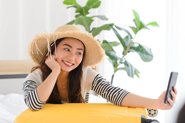 Молодая азиатка-блоггер улыбается летом в повседневной одежде и соломенной шляпе, разговаривая на facetime видео с помощью смартфона в спальне - Фото, изображение