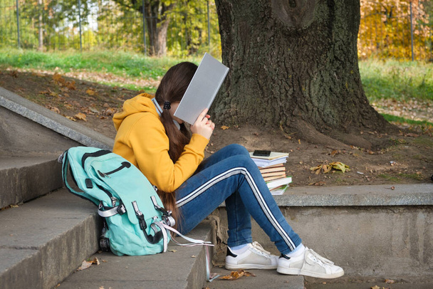 Moe tienermeisje leest een boek, bedekt haar gezicht met een boek zittend op de trap in het park. - Foto, afbeelding