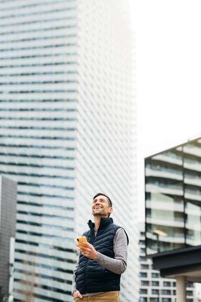 背景にスマートフォン、モダンな建物を使用して、カジュアルな服でフリーランスの男性。都市部での遠隔作業 - 写真・画像