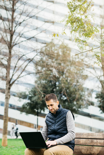 Freelancer mężczyzna w ubraniach casual, za pomocą smartfona, nowoczesny budynek na tle. Praca zdalna w środowisku miejskim - Zdjęcie, obraz