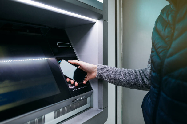 Main d'homme avec un smartphone utilisant un distributeur automatique de billets pour retirer de l'argent - Photo, image