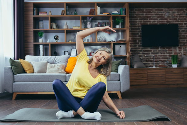 Senior gray-haired woman doing fitness at home, doing morning exercises on a sports mat sitting on the floor at home - Фото, изображение