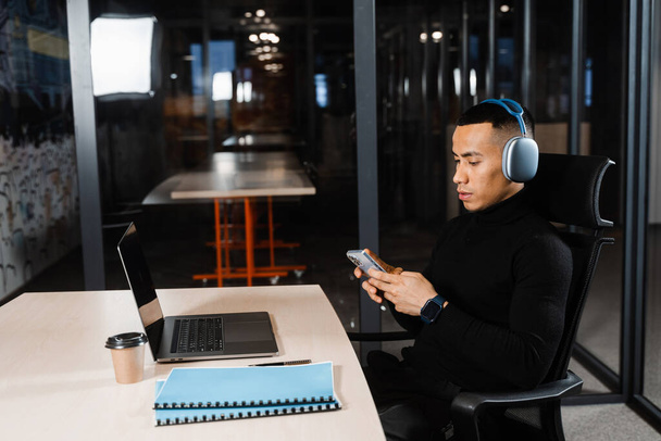 Asian man in headphones working online, listen to music and check his phone in office. Asian handsome guy with laptop in coworking space - Photo, Image