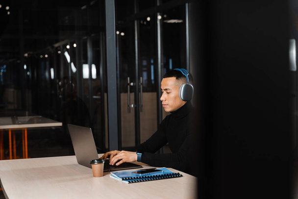 Азиатский мужчина в наушниках работает онлайн и слушает музыку в офисе. Азиатский красавчик с ноутбуком в коворкинге - Фото, изображение