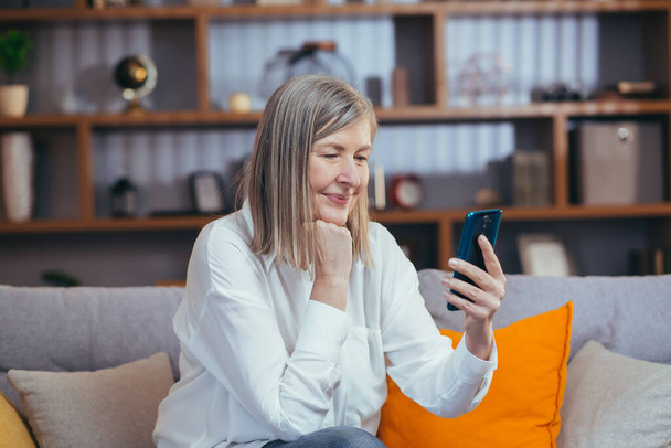 boldog idős nő használ telefont az online kommunikáció, ül a kanapén otthon este és mosolyog - Fotó, kép