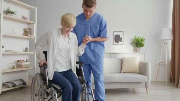 Hombre profesional terapeuta que ayuda a la mujer madura a levantarse de la silla de ruedas y caminar, poniéndose saludable después del accidente - Imágenes, Vídeo