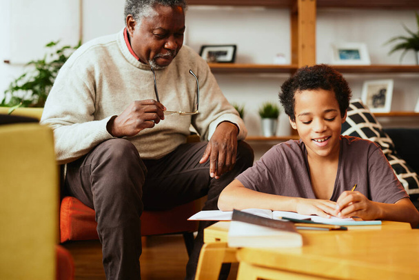 Um avô afro-americano está sentado em casa com seu neto e ajudá-lo com os trabalhos de casa. - Foto, Imagem