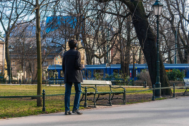 Lyhyttakkinen nuori mies pitää kädessään puhelinta, josta hän kuuntelee musiikkia langattomissa kuulokkeissa kävellessään kaupungissa alkukeväällä. - Valokuva, kuva