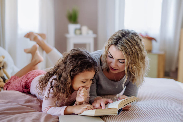 Happy mother with her little daughter lying on bed and reading book at home. - 写真・画像