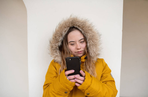 Happy young woman with Down syndrome weraing parka, leaning the wall and using smartphone. - Photo, Image