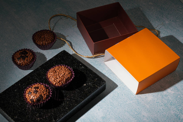 Low key photo. Brigadeiros arranged on a shiny black marble stone, next to a box with sisal rope_side view. - Foto, Imagen