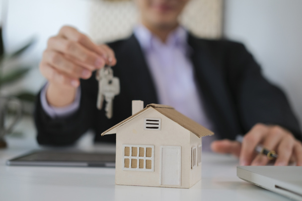Modello di piccola casa su tavolo bianco con agente immobiliare in possesso di chiavi in background. Concetto di investimento immobiliare. - Foto, immagini