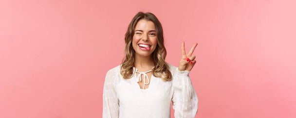 Close-up portrait of friendly joyful blond girl in white dress, sending positive vibes, smiling pleased and wink, stick tongue showing peace sign, looking kawaii over pink background - Фото, изображение