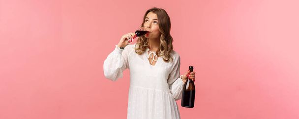 休日、春とパーティーのコンセプト。若いエレガントな幸せなブロンドの女性の肖像画、白のトレンディーなドレスを着て、喜んで見上げガラスからワインを飲んで、良い飲み物を試飲、瓶を保持 - 写真・画像