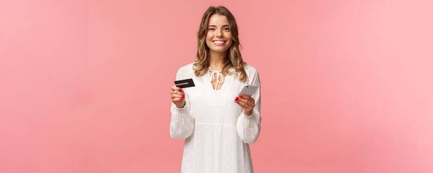 Portrait of happy smiling blond woman order food online, use application for shopping internet store, holding credit card and smartphone, grinning delighted at camera, pink background - Photo, image