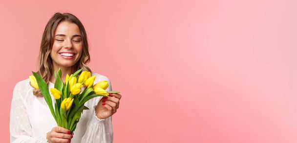 Frühling, Glück und Feiern. Nahaufnahme Porträt des schönen romantischen lächelnden Mädchens, das Blütenblatt der gelben Tulpe berührt, Blumen hält, Blumenstrauß auf Datum erhält, stehend rosa Hintergrund - Foto, Bild
