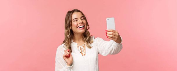 Zbliżenie portret wesołej, optymistycznej, uśmiechniętej blondynki, ubranej w białą sukienkę, śmiejącej się jako nagranie wideo, dzwoniącej przyjaciółki z aplikacji mobilnej, robiącej zdjęcia, selfie na smartfonie, różowe tło - Zdjęcie, obraz