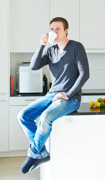Dieses Haus lebt von Lachen und viel starkem Kaffee. Porträt eines jungen Mannes, der auf seinem Küchentisch sitzt und Kaffee trinkt. - Foto, Bild