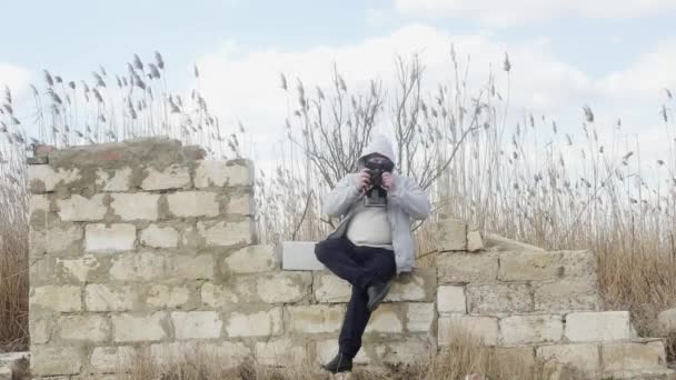  ένας άντρας με κουκούλα και μάσκα κάθεται σε έναν παλιό κατεστραμμένο τοίχο και τραβάει φωτογραφίες σε μια ρετρό κάμερα στο φόντο καλάμια. - Πλάνα, βίντεο