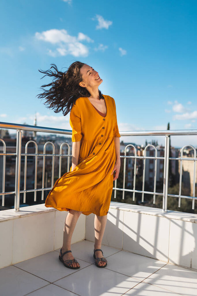 Dívka ve žlutých šatech stojí na balkóně během silného větru, usmívající se žena stojí na střeše s výhledem na město a vítr nafoukne její šaty, atraktivní dívka se dívá na město - Fotografie, Obrázek