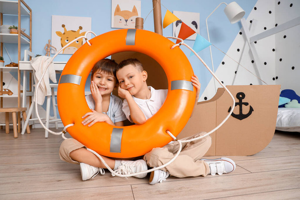 Vtipní malí kluci si hrají se záchrannou bójí a lepenkovou lodí doma - Fotografie, Obrázek
