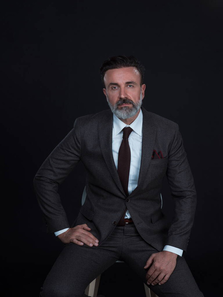 Portrait of adult businessman wearing trendy suit and sitting in modern studio on stylish chair against the black background - Foto, Bild
