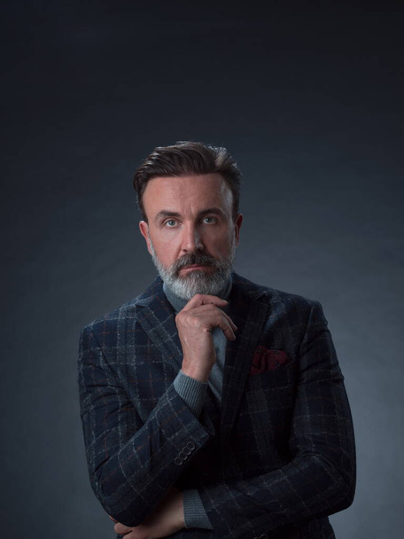Portrait of a stylish elegant senior businessman with a beard and casual business clothes in photo studio isolated on dark background gesturing with hands - Фото, изображение