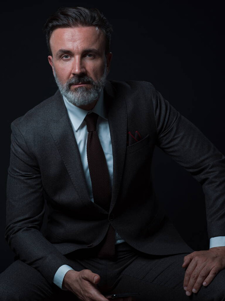 Portrait of adult businessman wearing trendy suit and sitting in modern studio on stylish chair against the black background - Φωτογραφία, εικόνα