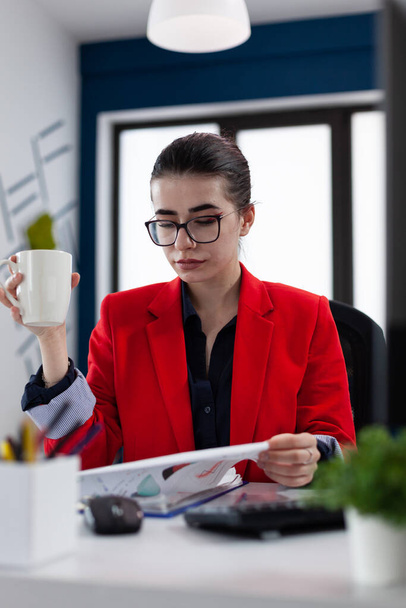 Businesswoman with glasses holding cup and looking at clipboard with chart. - Φωτογραφία, εικόνα