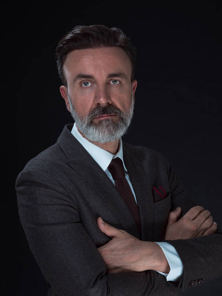 Portrait of a stylish elegant senior businessman with a beard and casual business clothes in photo studio isolated on dark background gesturing with hands - Foto, Imagem