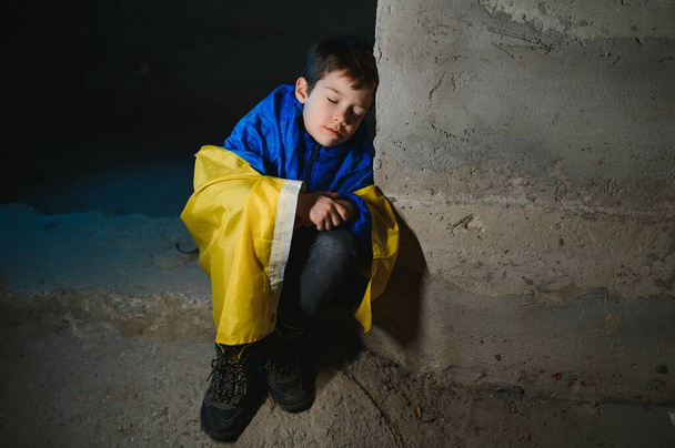 A sad Ukrainian boy wrapped in a Ukrainian flag, hiding from bombings in a bomb shelter in the city of Mariupol or Kiev. Russia's aggression. The war in Ukraine and 2022. - 写真・画像