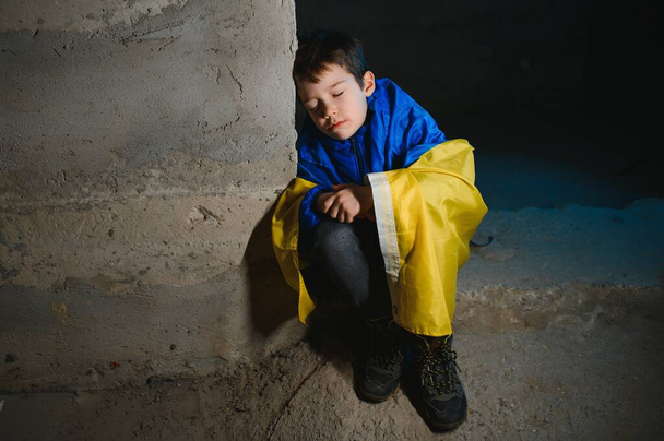 Egy szomorú ukrán fiú, ukrán zászlóba csomagolva, bombázások elől bujkálva egy óvóhelyen Mariupol városában vagy Kijevben. Oroszország agressziója. A háború Ukrajnában és 2022. - Fotó, kép