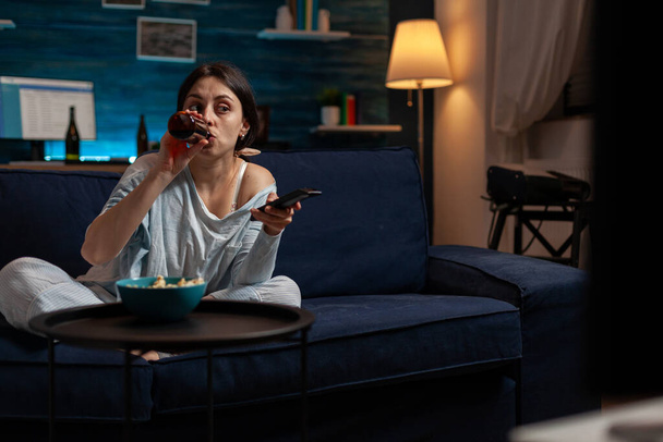 Young woman drinking beer from bottle and switching TV channels - Фото, изображение