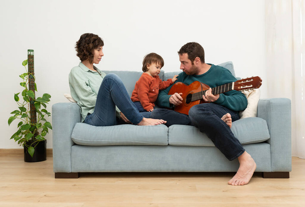 Eltern und Kleinkind sitzen an einem Tisch und haben Zeit miteinander. Mann spielt eine spanische Gitarre, während sein kleines Mädchen und seine Frau ihn anschauen und die Musik genießen.  - Foto, Bild