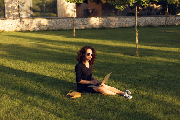 Attractive young woman in sunglasses sitting on lawn with legs during summer day while using laptop. Working outside. - Photo, Image