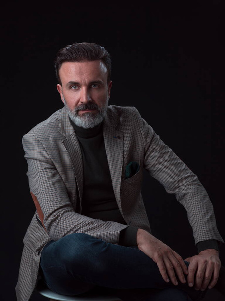 Portrait of adult businessman wearing trendy suit and sitting in modern studio on stylish chair against the black background - Fotografie, Obrázek