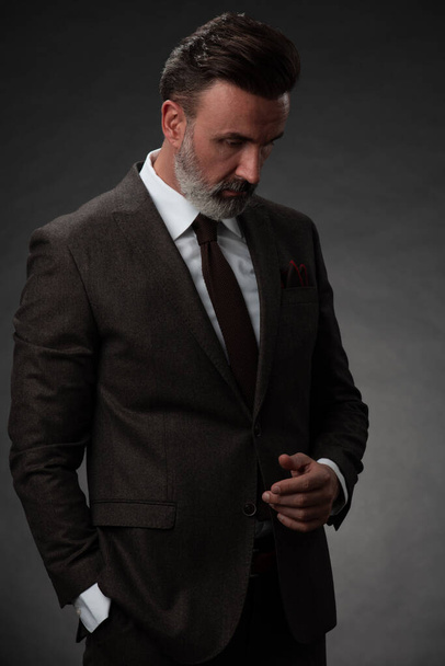 Retrato de um elegante homem de negócios sênior elegante com barba e roupas de negócios casuais em estúdio de fotografia isolado em fundo escuro gesticulando com as mãos - Foto, Imagem
