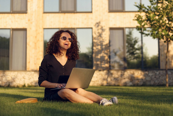 Porträt einer fröhlichen jungen Frau mit Sonnenbrille, die mit überkreuzten Beinen auf grünem Gras sitzt und mit Laptop lächelt. - Foto, Bild